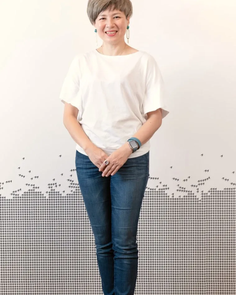 代表の髙橋元子。白いTシャツにジーンズで、サロンの壁の前に立っている